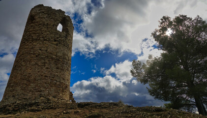 Arab tower of Albolote seen from below next to a tree that lets through solar rays and a dramatic sky in the background