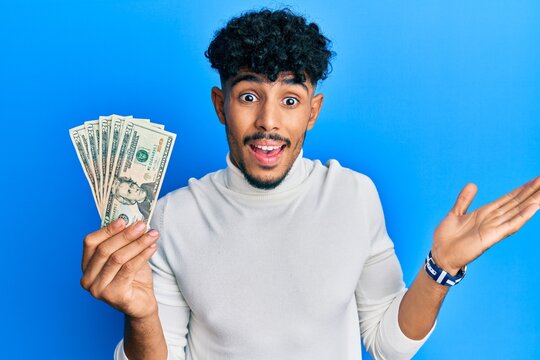 Young arab handsome man holding 20 dollars banknotes celebrating achievement with happy smile and winner expression with raised hand