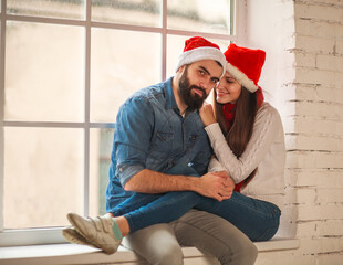 Young merry Christmas couple in Santa hats sitting on windowsill at home happily together