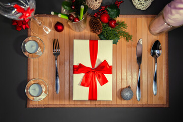 Table setting. А Gift box with red satin ribbon on a wooden napkin with a knife, spoon and fork near Christmas decorations and candles. Black background.