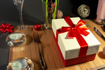 Table setting. А Gift box with red satin ribbon on a wooden napkin with a knife, spoon and fork near Christmas decorations and candles. Black background.