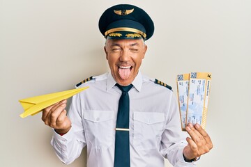 Handsome middle age mature pilot man holding paper plane and boarding pass sticking tongue out...