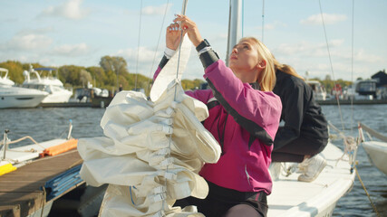 A young blonde with a teammate puts the mainsail on the afterstay of her sports yacht. Straightens the sails and prepares the rigging for the race.