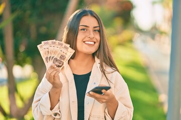 Young hispanic girl smiling happy using smartphone and hoding mexican pesos banknotes at the park. - obrazy, fototapety, plakaty