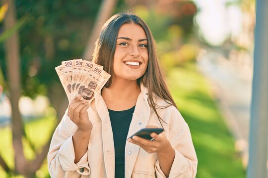 Young hispanic girl smiling happy using smartphone and hoding mexican pesos banknotes at the park.