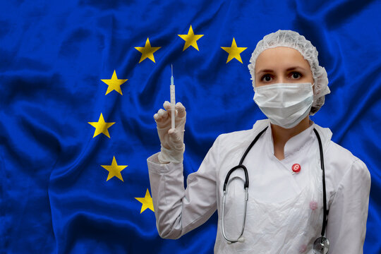 Young woman doctor in medical uniform on the background of the flag of European Union is holding a syringe. Concept of new coronavirus vaccine, coronavirus COVID-19 vaccination