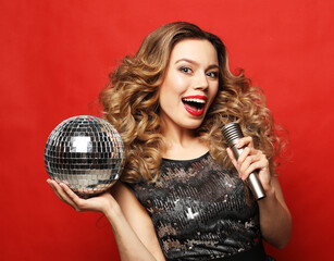 Young blond woman with long wavy hair dressed in evening dress holding a microphone and disco ball, singing and smiling