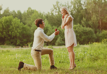 Fototapeta premium Happy couple, man kneeling down and proposing a ring to his woman outdoors on the grass, wedding concept