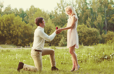 Happy couple, man kneeling down and proposing a ring to his woman outdoors on the grass, wedding concept