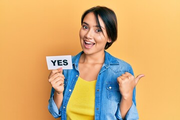 Beautiful young woman with short hair holding yes reminder pointing thumb up to the side smiling happy with open mouth