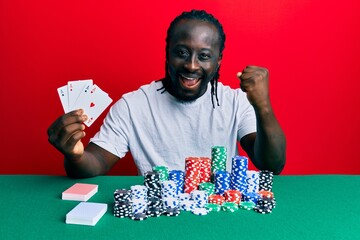 Handsome young black man playing poker holding cards screaming proud, celebrating victory and success very excited with raised arms