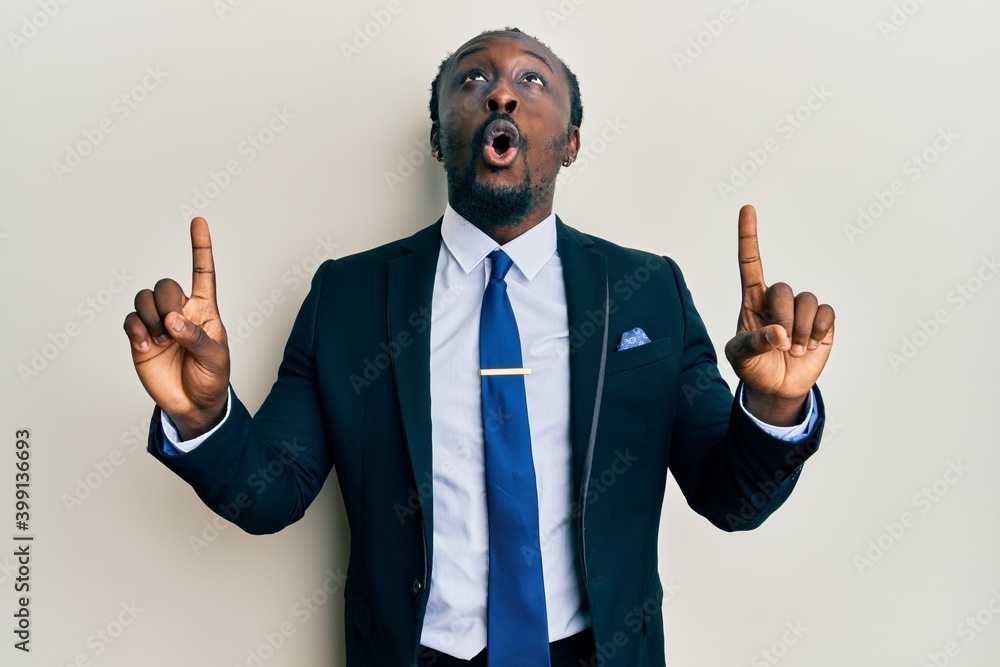 Wall mural Handsome young black man wearing business suit and tie amazed and surprised looking up and pointing with fingers and raised arms.