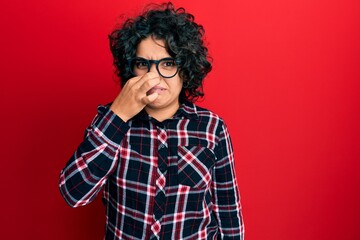Young hispanic woman with curly hair wearing casual clothes and glasses smelling something stinky...