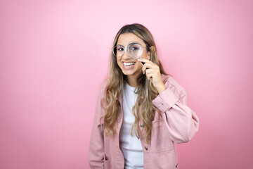 Young beautiful blonde woman with long hair standing over pink background surprised looking through a magnifying glass