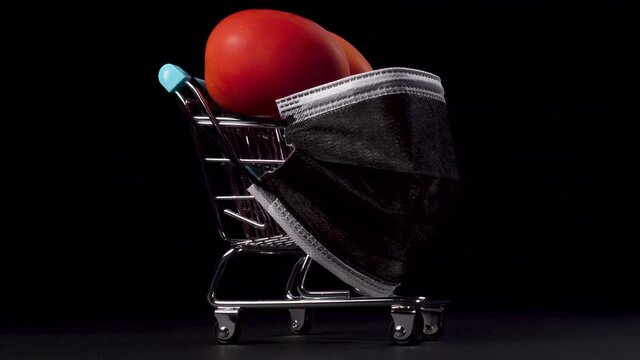 Mini supermarket trolley with medical protective mask and loaded red vegetables on a black background. Ripe tomatoes in a shopping cart. Safe online shopping and home delivery concept