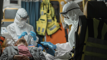 Paramedic in Coveralls Using Tablet Computer to Check the Diagnosis for the Injured Patient on the Way to Hospital. Emergency Medical Care Assistants Puts a Ventilation Mask in an Ambulance.