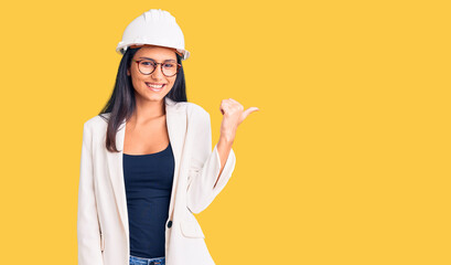 Young beautiful latin girl wearing architect hardhat and glasses smiling with happy face looking and pointing to the side with thumb up.