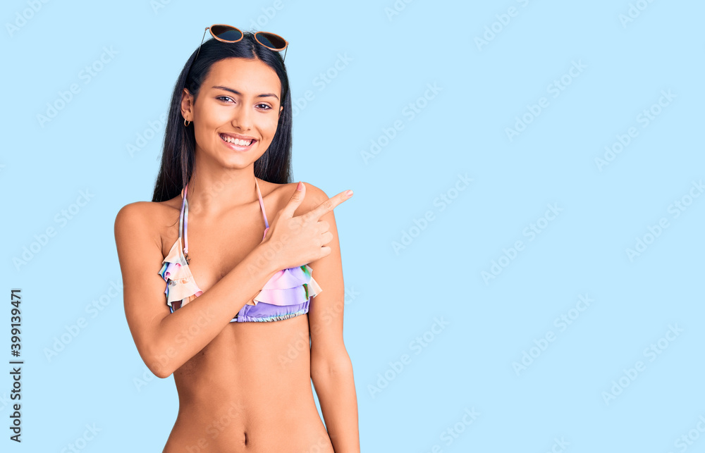 Poster Young beautiful latin girl wearing bikini and sunglasses cheerful with a smile of face pointing with hand and finger up to the side with happy and natural expression on face