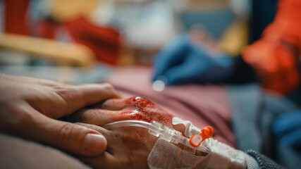 Close Up Shot of Professional Paramedic Providing Medical Help to an Injured Patient on the Way to Healthcare Hospital. Shot is Focused on Wounded Hand with Blood and an Intravenous Line in Wrist.