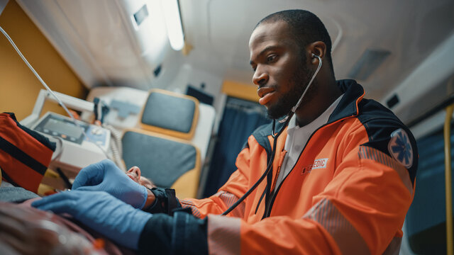 Close Up Portrait Shot of an African American Professional Paramedic Providing Medical Help to an Injured Patient on the Way to Hospital. Emergency Care Assistant Using Stethoscope in an Ambulance.