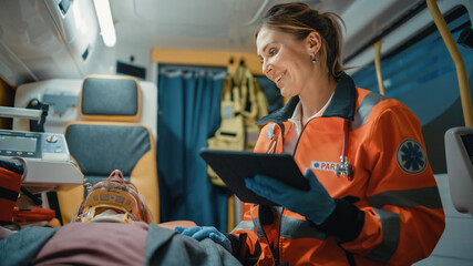 Female EMS Professional Paramedic Using Tablet Computer to Fill a Questionnaire for the Injured Patient on the Way to Hospital. Emergency Care Assistant Comforting the Patient in an Ambulance. - obrazy, fototapety, plakaty