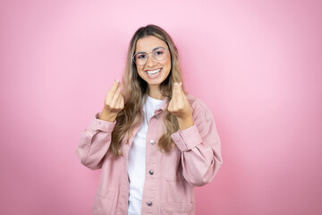Young beautiful blonde woman with long hair standing over pink background doing money gesture with...