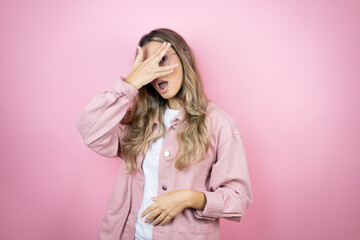 Young beautiful blonde woman with long hair standing over pink background peeking in shock covering face and eyes with hand, looking through fingers with embarrassed expression