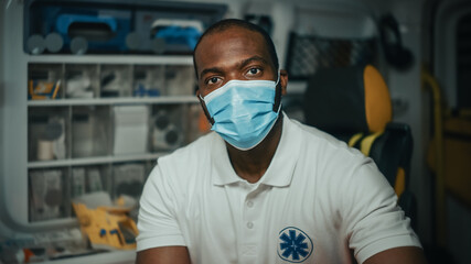Fototapeta na wymiar Portrait of a Black African American EMS Paramedic Looks at Camera While Wearing a Safety Face Mask in Ambulance Vehicle. Emergency Medical Technician Outside the Hospital. Covid-19 Concept.