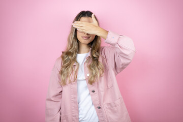 Young beautiful blonde woman with long hair standing over pink background serious and covering her eyes with her hand