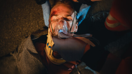 Portrait of a Young Injured Man Involved in a Traffic Accident is Being Saved by a Medical Team of EMS Paramedics on the Street at Night. Emergency Care Assistants Use a Ventilation Mask.