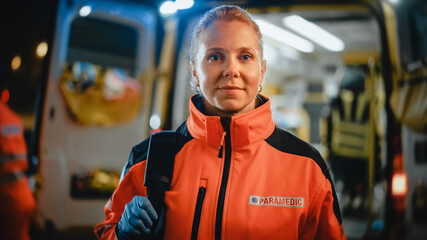 Portrait of a Female EMS Paramedic Proudly Standing in Front of Camera in High Visibility Medical Orange Uniform with "Paramedic" Text Logo. Successful Emergency Medical Technician or Doctor at Work. © Gorodenkoff