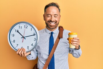 Handsome middle age business man drinking a cup of coffee and holding big clock smiling and...