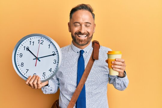 Handsome middle age business man drinking a cup of coffee and holding big clock smiling and laughing hard out loud because funny crazy joke.