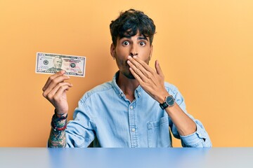 Young hispanic man holding 50 dollar banknote sitting on the table covering mouth with hand, shocked and afraid for mistake. surprised expression