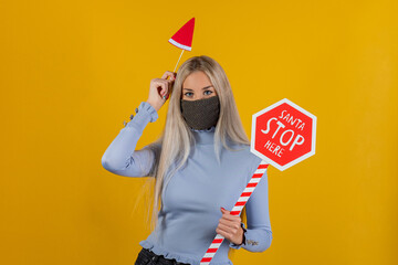 Woman in protective face mask holding with hand santa claus hat and road sign on yellow background. Party image in pandemic period. Playful young girl celebrating and joking in covid19 time