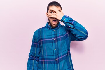 Young handsome man wearing casual shirt peeking in shock covering face and eyes with hand, looking through fingers afraid