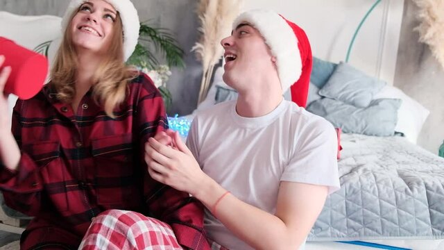 Young happy couple dancing and dabbles on the floor near the bed wearing santa hats and pajamas giving gifts for Christmas