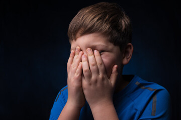 white teenage boy with light brown hair in a blue sports t-shirt covering his face with two hands on  isolated background