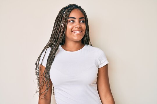 Young african american woman with braids wearing casual white tshirt looking away to side with smile on face, natural expression. laughing confident.