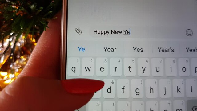 Female hand is typing with T9 a message Happy New Yearon on the screen of a white smartphone. Background of golden tinsel.