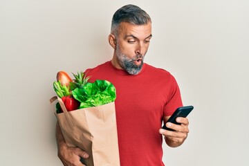 Middle age handsome man holding bag of groceries using smartphone in shock face, looking skeptical...