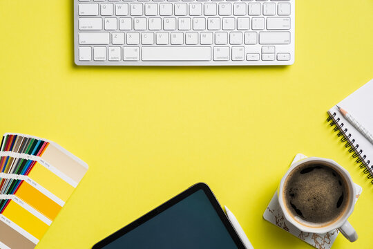 Professional creative graphic designer desk with computer keyboard, tablet, color palette, cup of coffee, notebook on yellow background. Flat lay, top view.