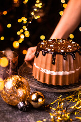 Holding tasty Three Chocolates mousse cake with ganache in Christmas and New Year decorations. Chocolate cake with layers. Bokeh lights background. Vertical orientation. Christmas Eve. Low Key.