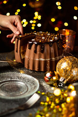 Cutting tasty Three Chocolates mousse cake with ganache in Christmas and New Year decorations. Chocolate cake with layers. Bokeh lights background. Vertical orientation. Christmas Eve. Low Key.
