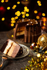 A piece of tasty Three Chocolates mousse cake with ganache in Christmas and New Year decorations. Chocolate cake with layers. Bokeh lights background. Vertical orientation. Christmas Eve. Low Key.