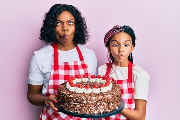 Beautiful african american mother and daughter wearing baker apron holding homemade cake making fish face with mouth and squinting eyes, crazy and comical.