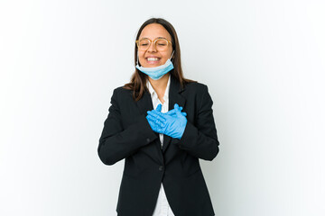 Young business latin woman wearing a mask to protect from covid isolated on white background laughing keeping hands on heart, concept of happiness.