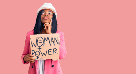 Young african american woman wearing architect helmet holding woman power banner serious face thinking about question with hand on chin, thoughtful about confusing idea