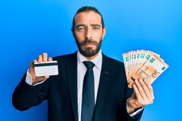 Attractive business man with long hair and beard holding credit card and euros relaxed with serious...