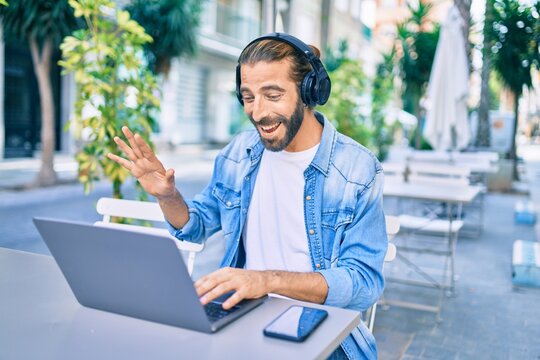 Young middle eastern man doing video call using laptop and headphones at coffee shop terrace.